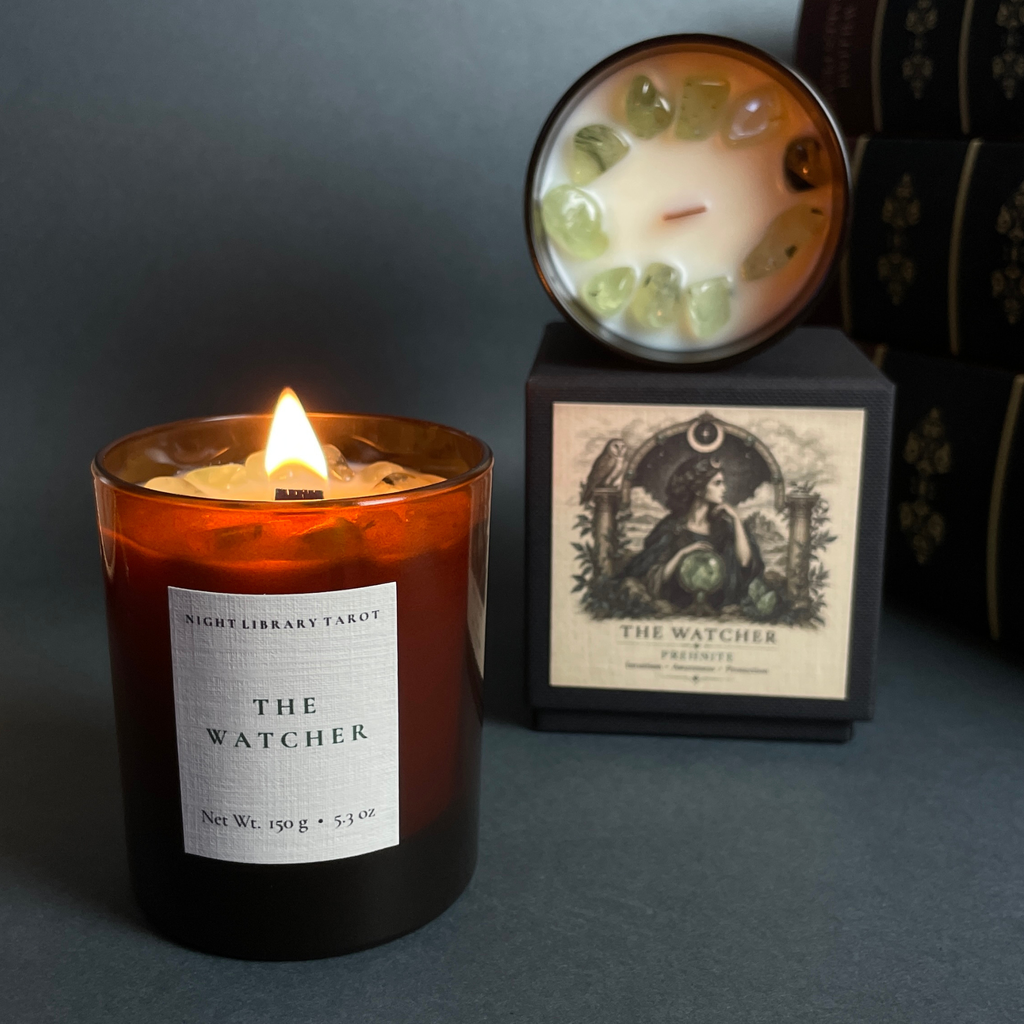 A lit amber glass jar candle labeled 'THE WATCHER' by Night Library Tarot sits in front of its black box packaging, which has a matching candle resting on top showing green crystal accents.