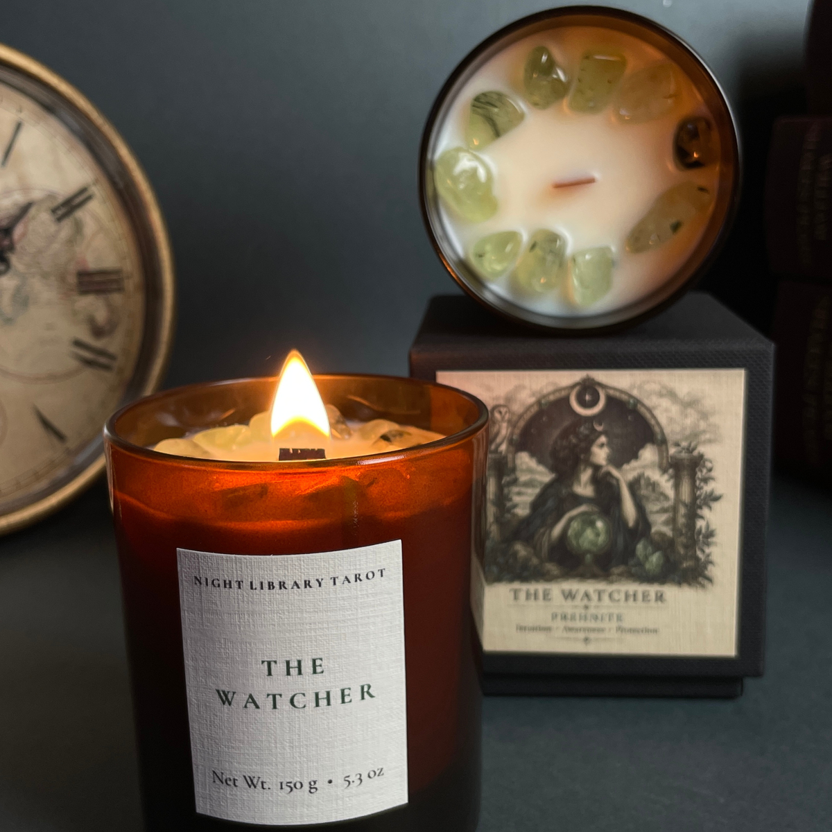 A lit amber glass candle labeled 'THE WATCHER' by Night Library Tarot, shown in the foreground with its black box and lid placed behind it on a dark tabletop. The lid reveals pale wax topped with translucent green crystal-like pieces.