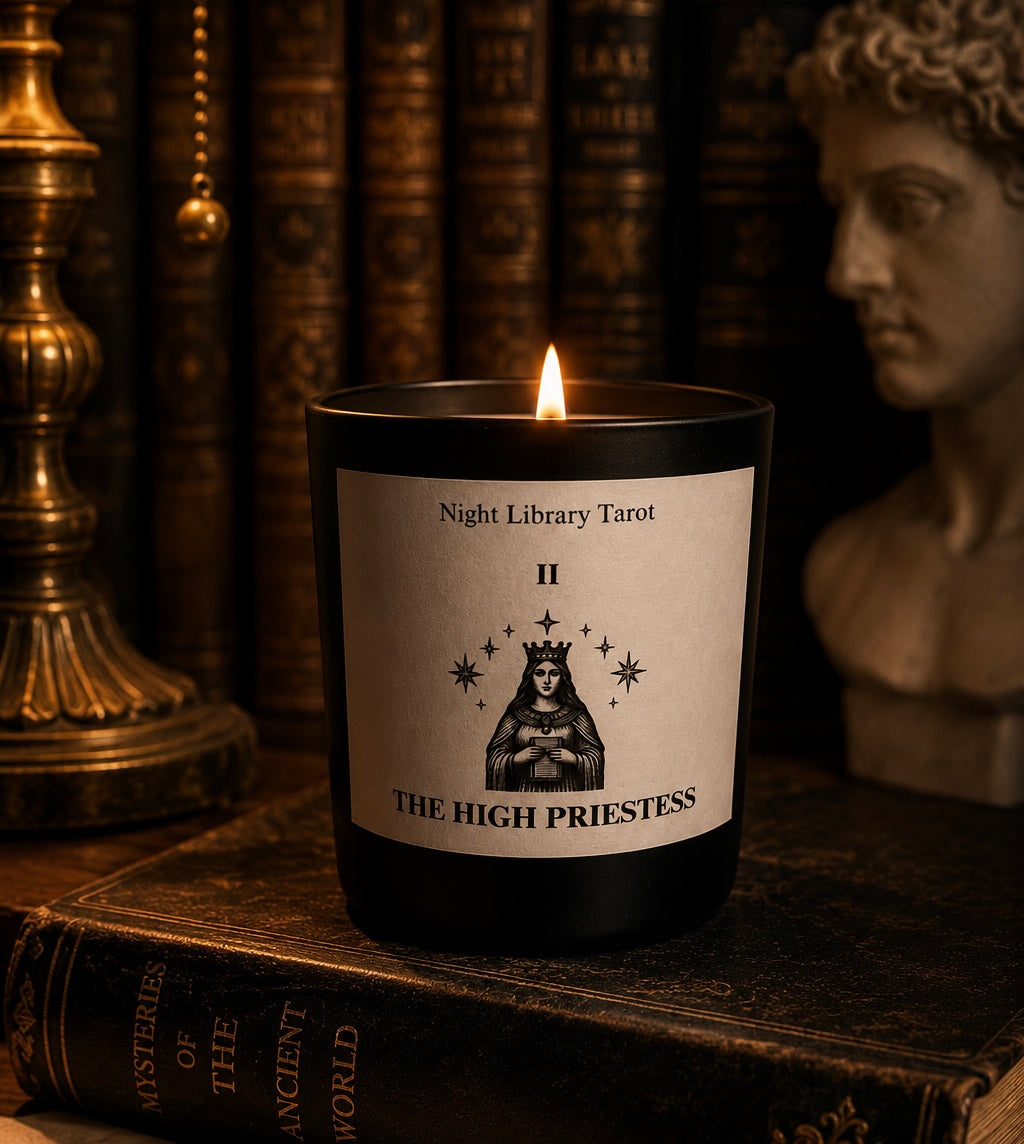 Candle labeled 'The High Priestess' from 'Night Library Tarot' on an old book with a statue and lamp in the background. Tarot inspired luxury candle. Dark Academia.