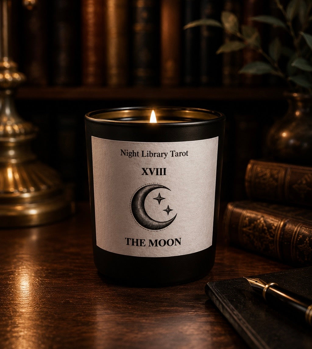 Candle labeled 'The Moon' from 'Night Library Tarot' on a wooden surface with books and a lamp in the background. The moon tarot inspired luxury candle. Dark academia.